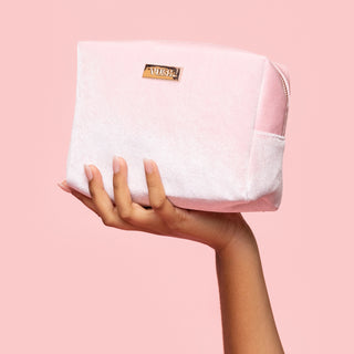 Hand holding pink soft cosmetics bag made of fuzzy velvet-like material with rose gold plaque that reads "VUSH" up against light pink background