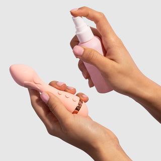 Left hand holding VUSH Rose 2 Precision Bullet Vibrator with rose detailing on head. Right hand holding VUSH Clean Queen Intimate Accessory Spray and spraying product onto vibrator.