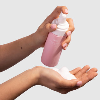Left hand holding 150ml pink bottle with white pump lid and white text that reads "VUSH IT'S ALL GOOD INTIMATE WASH" being pumped into right hand with white foam in it, against light grey background