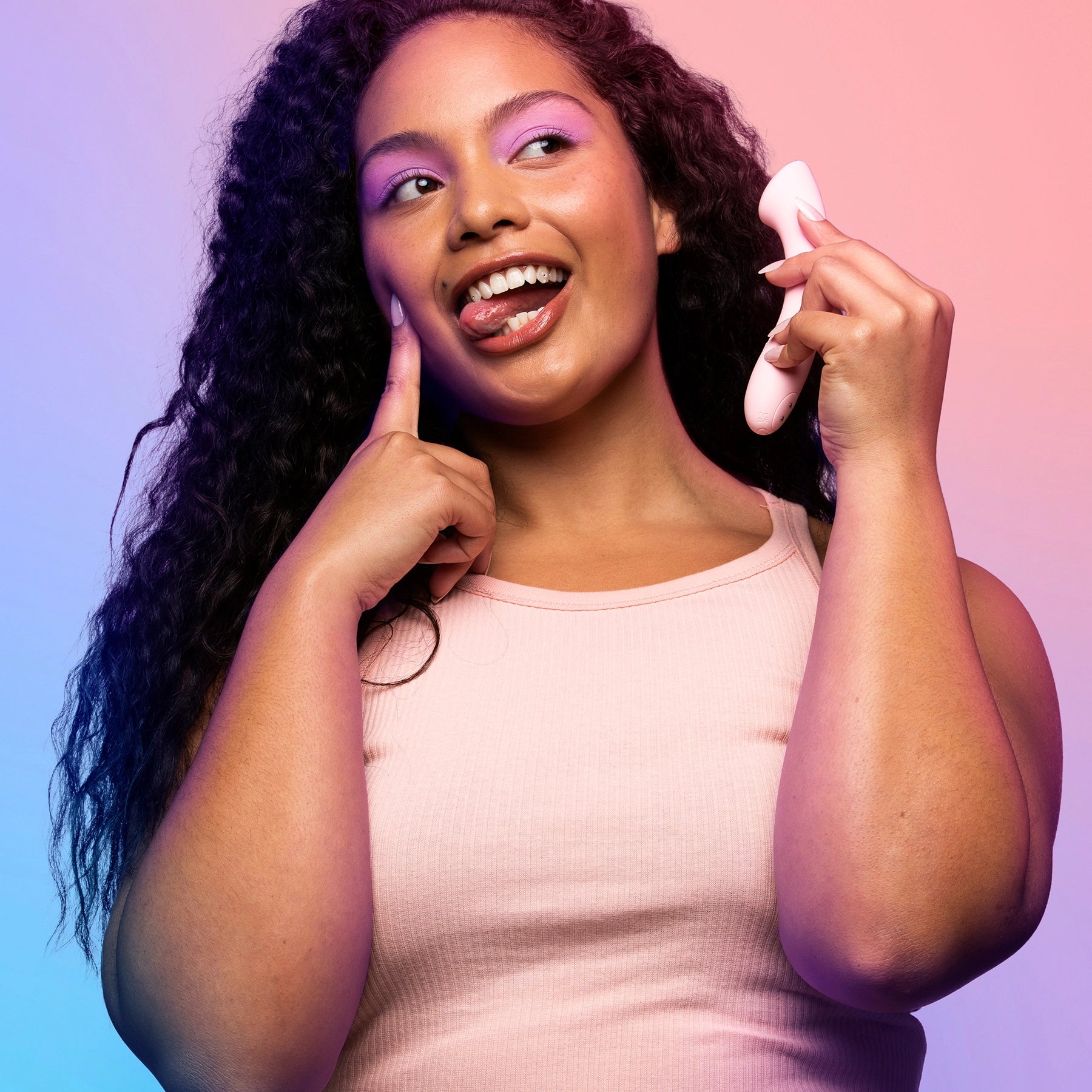 Young woman with long curly dark hair wearing baby pink tank top smiling and poking tongue out while holding VUSH Shine G Spot Vibrator against ear like a telephone