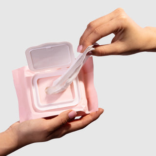 One hand holding pink packet with white lid while another hand pulls out white wipe from the packet